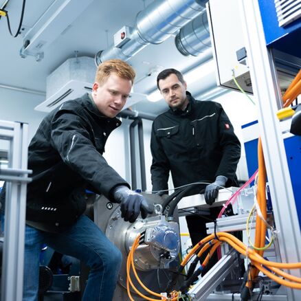 Zwei Mitarbeiter in kompletter Werkstattbekleidung arbeiten an einem orangen HV Kabel